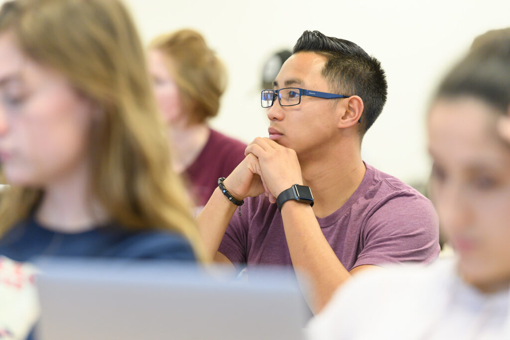 A student sits focused in a lecture with hands in a thinking position