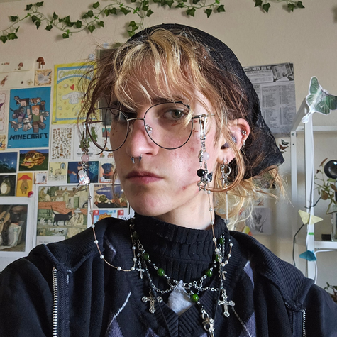 Lyd Paulsen is depicted shoulders up with a neutral expression on their face and their head angled to the left. They are a white nonbinary person with brown and blonde split dye bangs, wearing circular glasses and a black bandana and turtleneck.