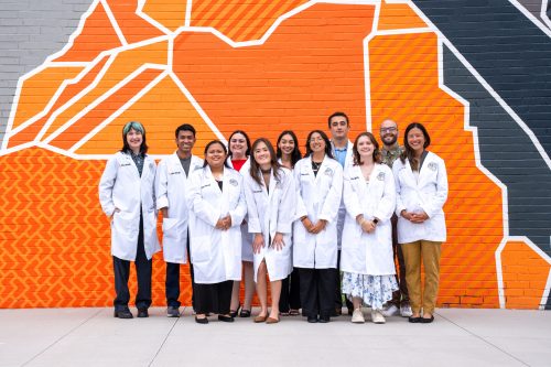 11 students at CSU-Spur pose for a group photo in their white lab coats in front of a wall portrait of orange and grey mountains