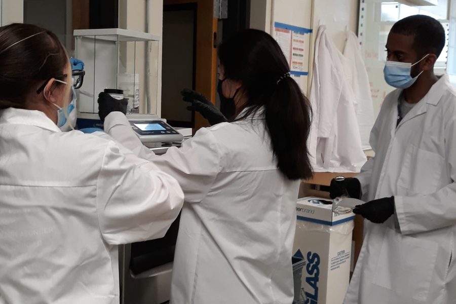 three students wearing white lab coats use lab equipment in the oura lab